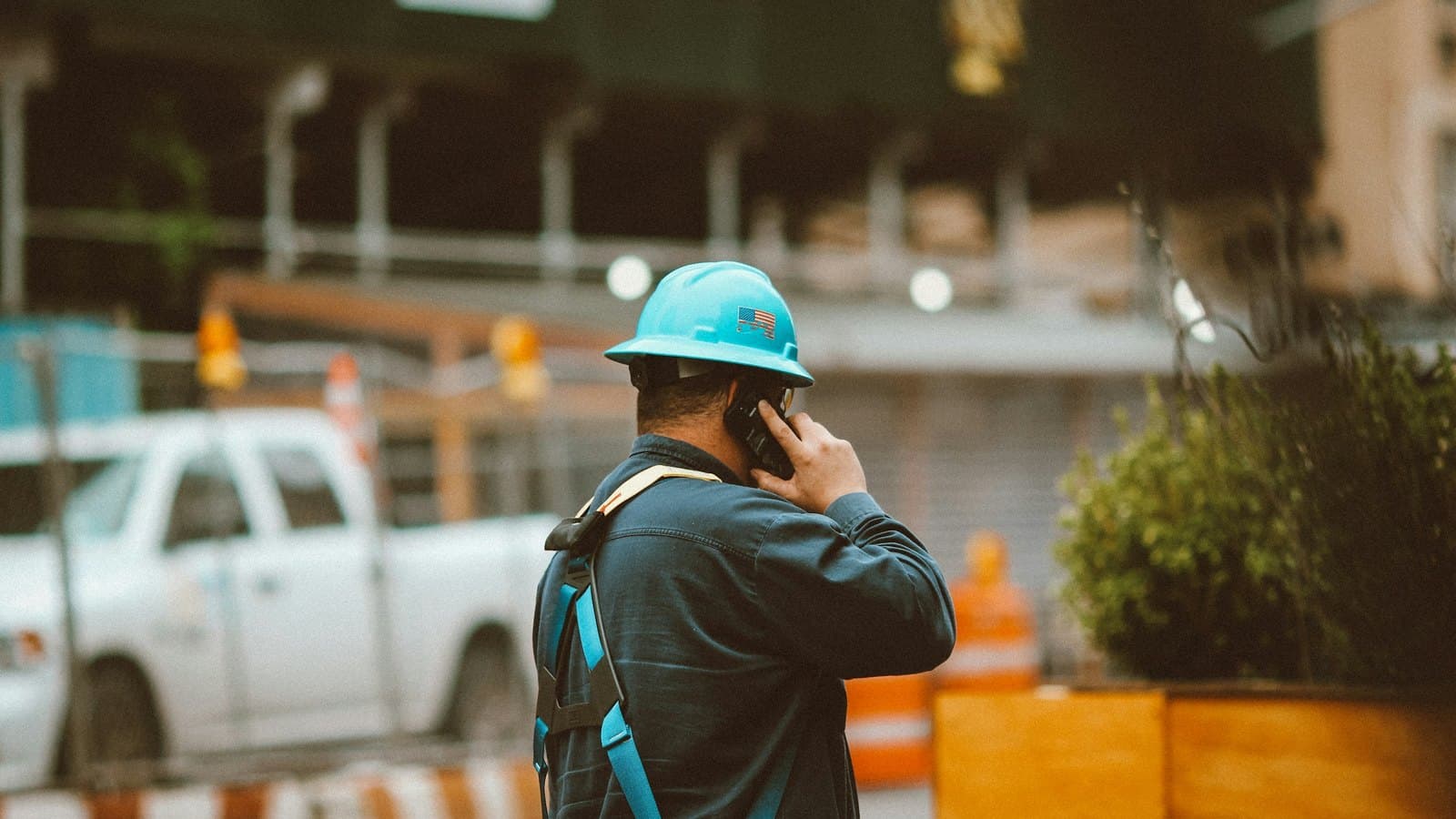 A worker in a hard hat on a city jobsite taking a call beside a pickup and scaffolding.