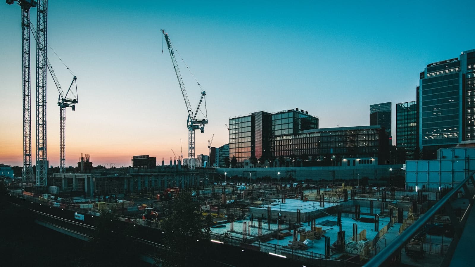 A wide dusk view over an active city jobsite: tower cranes above an excavated foundation of formwork and rebar, with lit office towers behind.